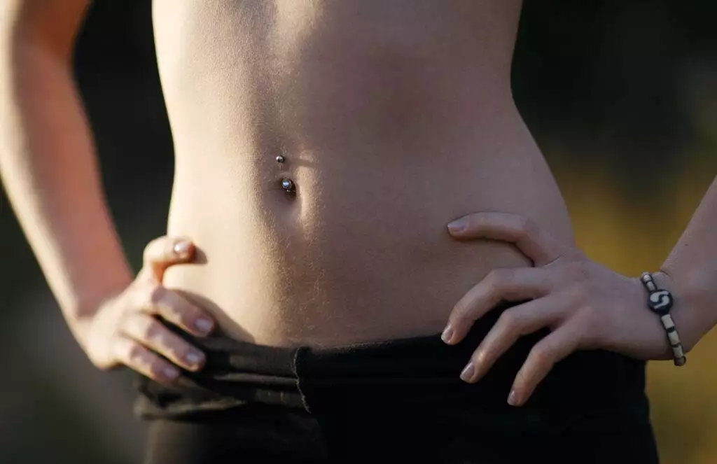 Close-up of a person's lower abdomen showcasing one of the hottest piercing trends—a navel piercing—while wearing black pants and a bracelet, with hands resting on hips.